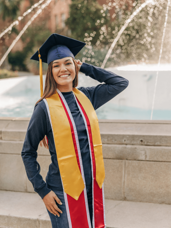 A young woman beaming with joy, wearing a graduation gown and cap, adorned with a gold graduation stole, celebrating her achievement after the ceremony.