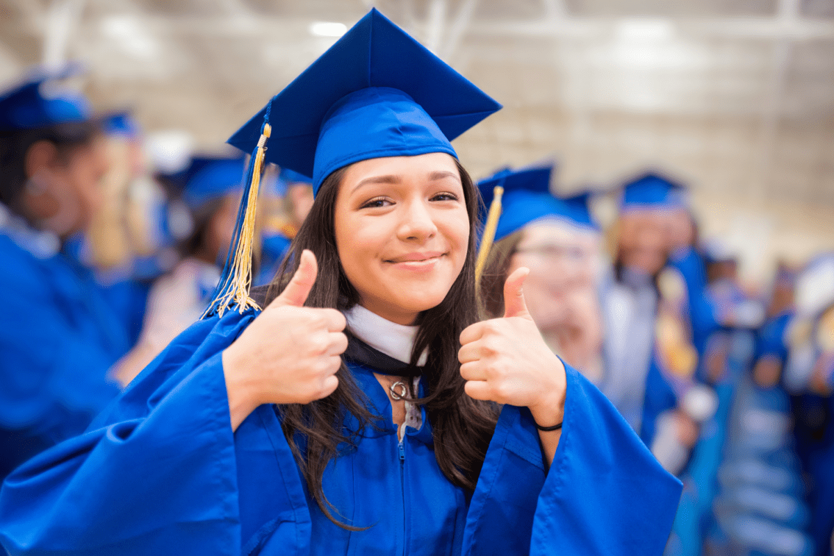 A young woman in a graduation cap and gown, smiling broadly and giving two thumbs up, celebrating her academic achievement with joy and pride.
